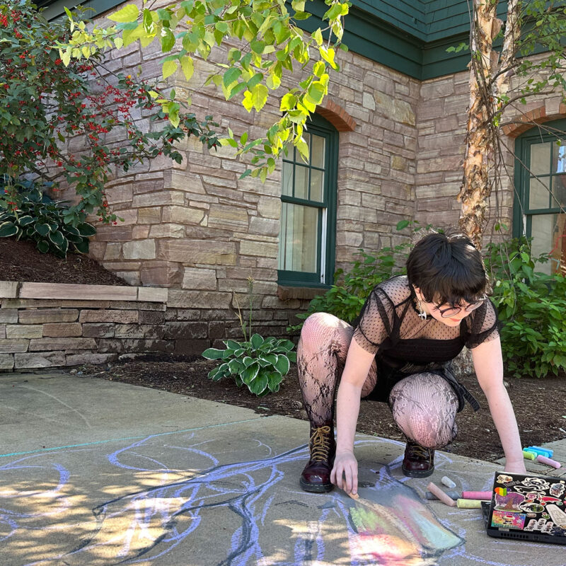 a student draws with chalk on the ground in the courtyard on a sunny day. they are looking at their laptop for art inspiration.