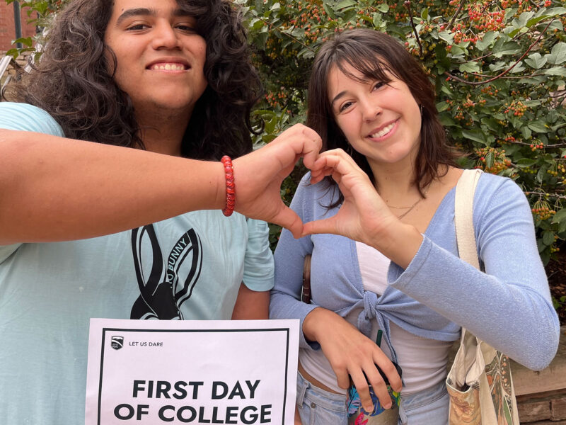 two students make a heart with their joining hands and hold up a sign that says "first day of school"