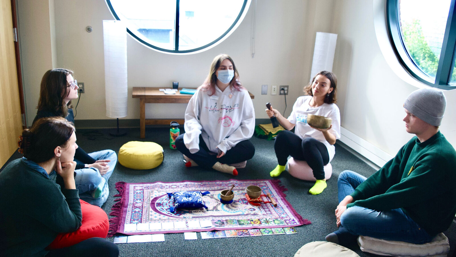 five students sit on the floor in the interfaith room getting ready to meditate. there are colorful pillows and blankets spread on the floor.