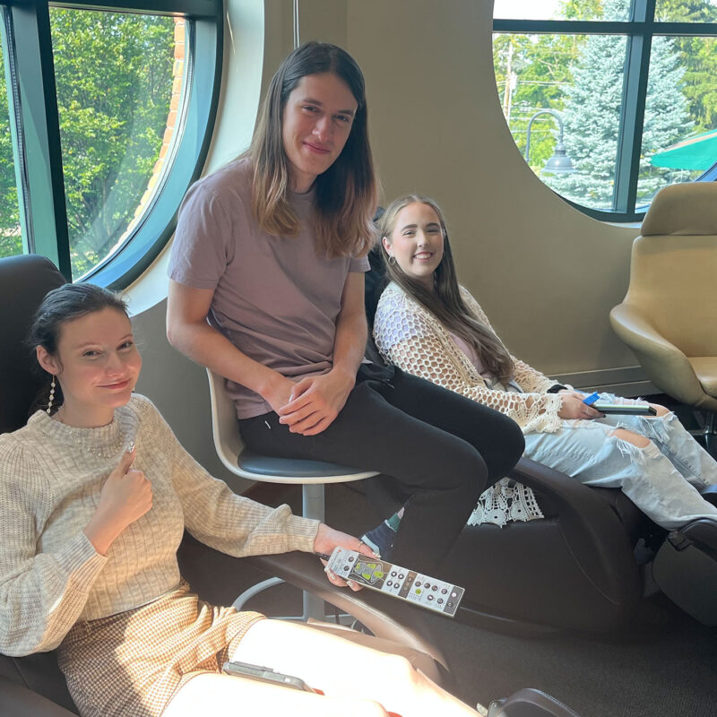 two students lounge on massage chairs in the smart space, while a third student sits between them, all smiling at the camera