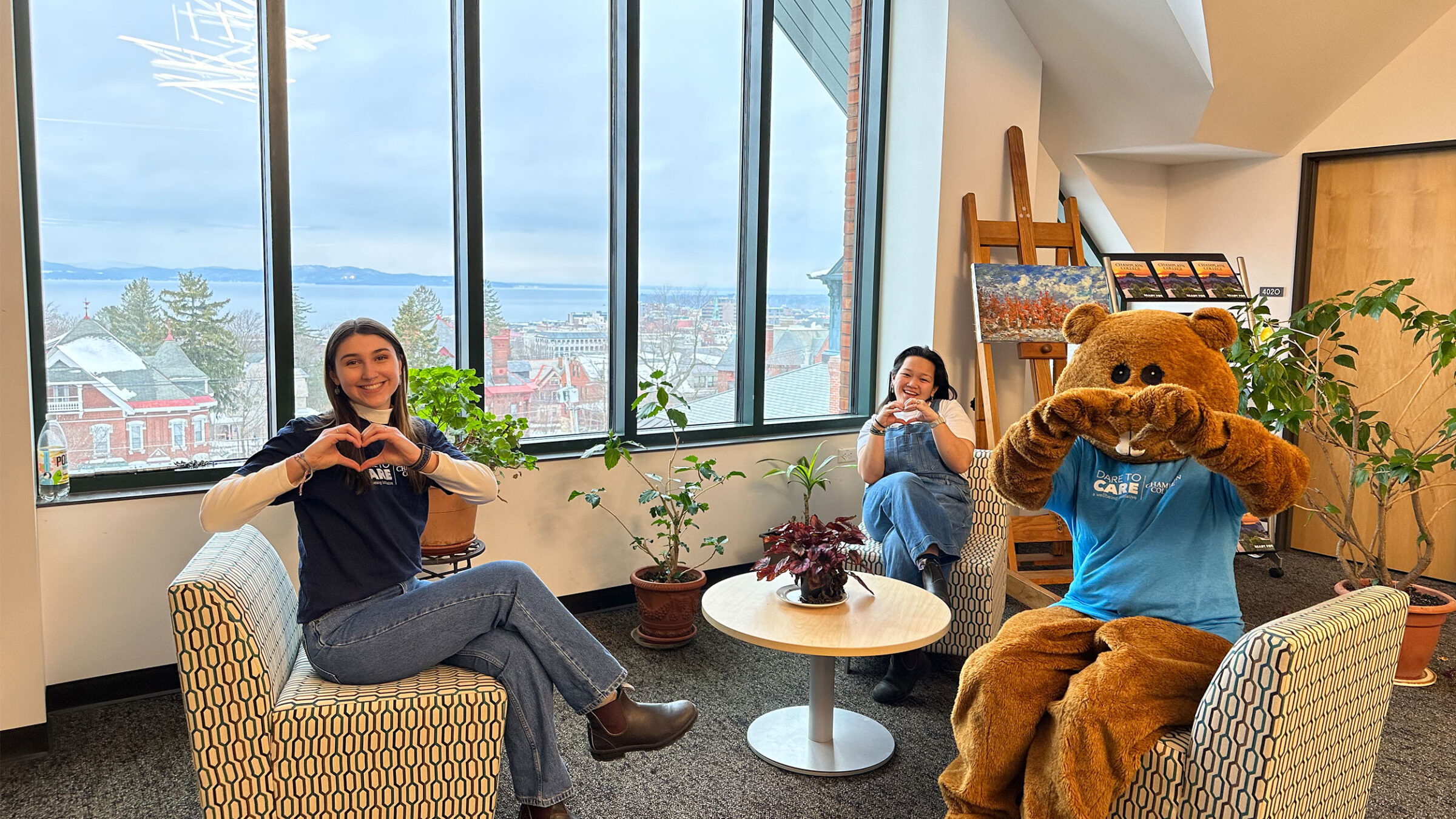 two students and chauncey the beaver mascot sit on chairs, making heart shapes with their hands. Lake Champlain is visible through the large windows behind them.