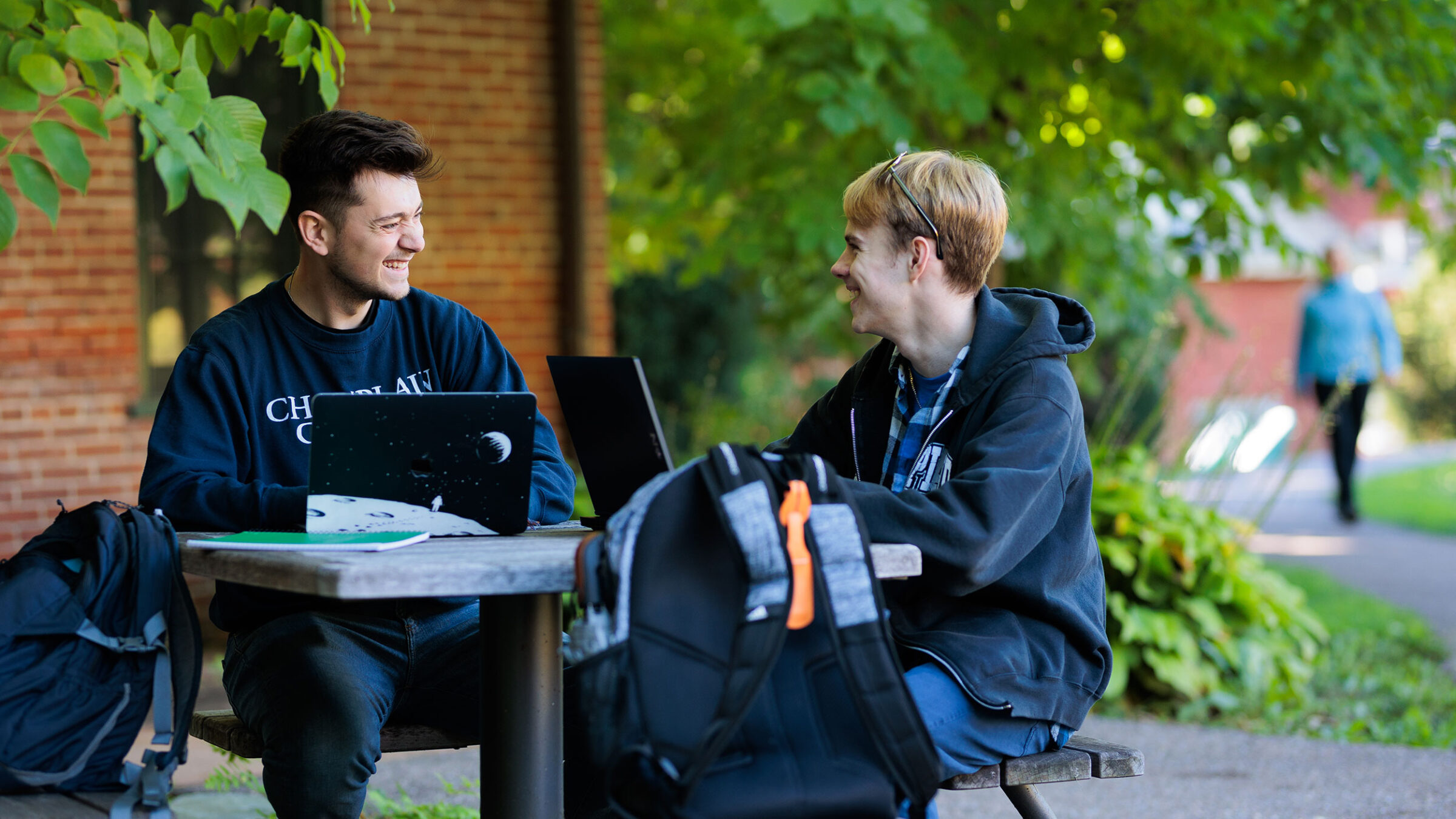 two students sit at a table in the green courtyard with their laptops out