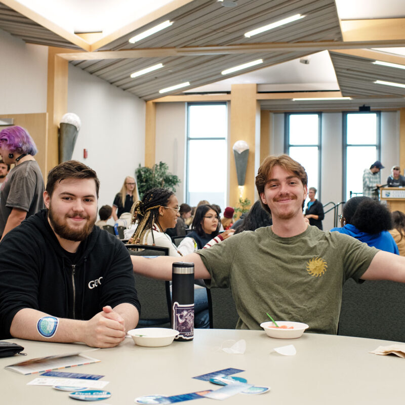 four students sit at a table in the champlain room, smiling at the camera with a crowded room seated behind them.