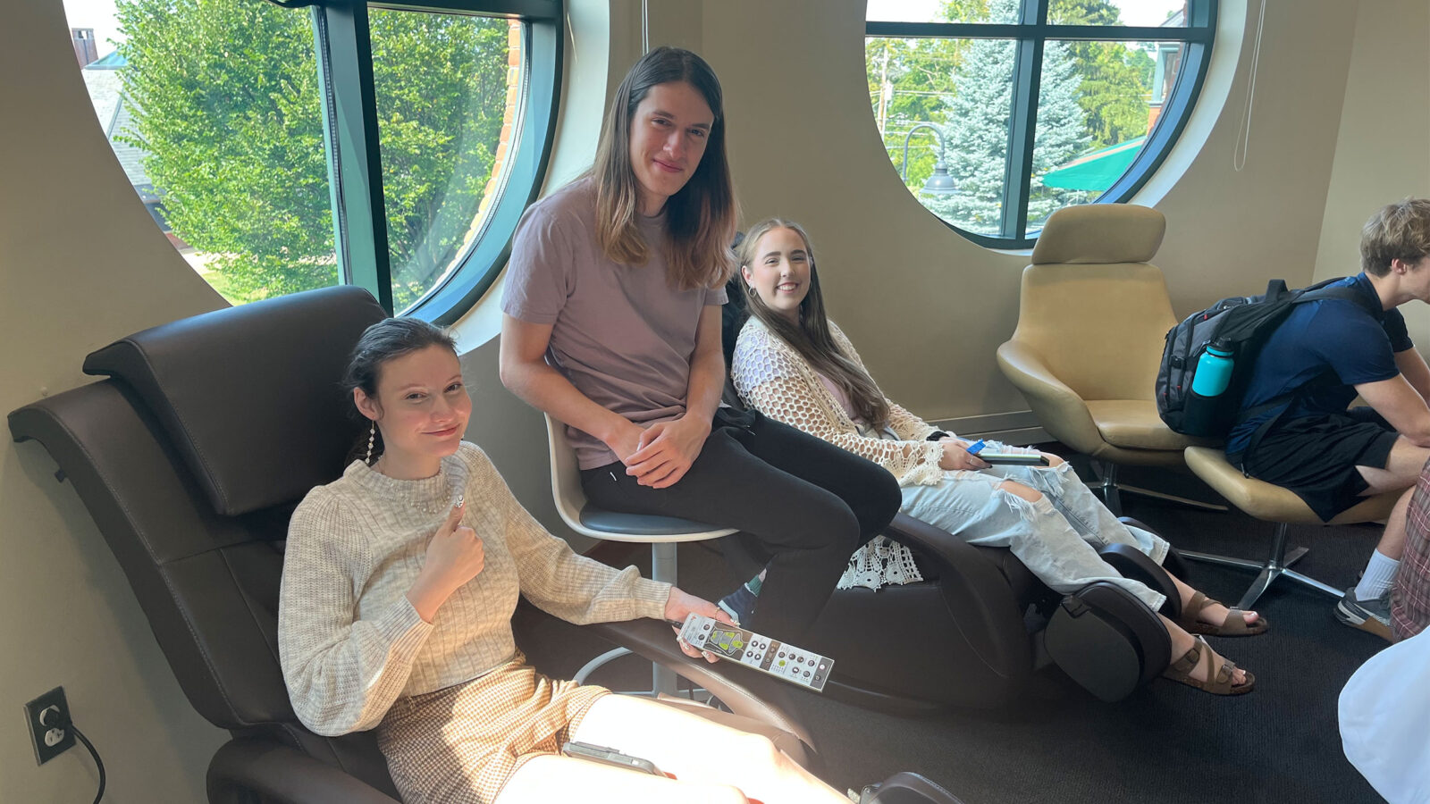 two students lounge on massage chairs in the smart space, while a third student sits between them, all smiling at the camera