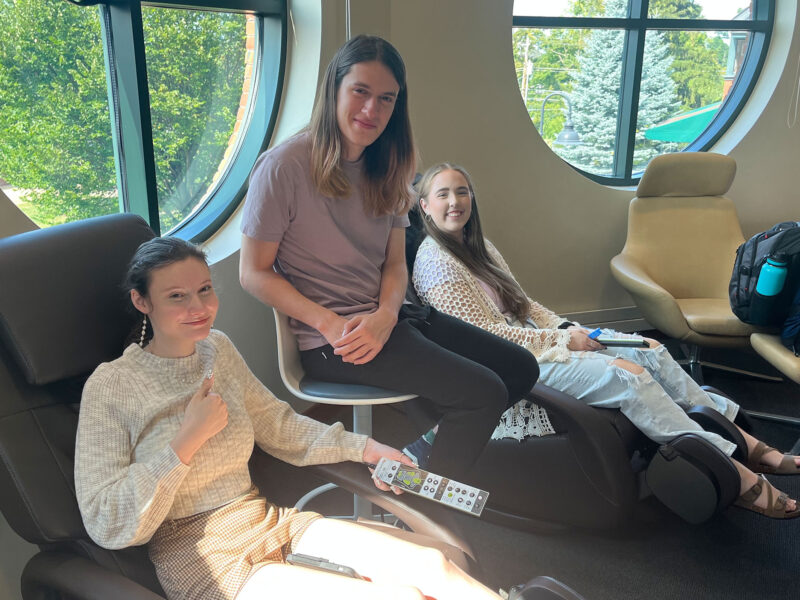 two students lounge on massage chairs in the smart space, while a third student sits between them, all smiling at the camera