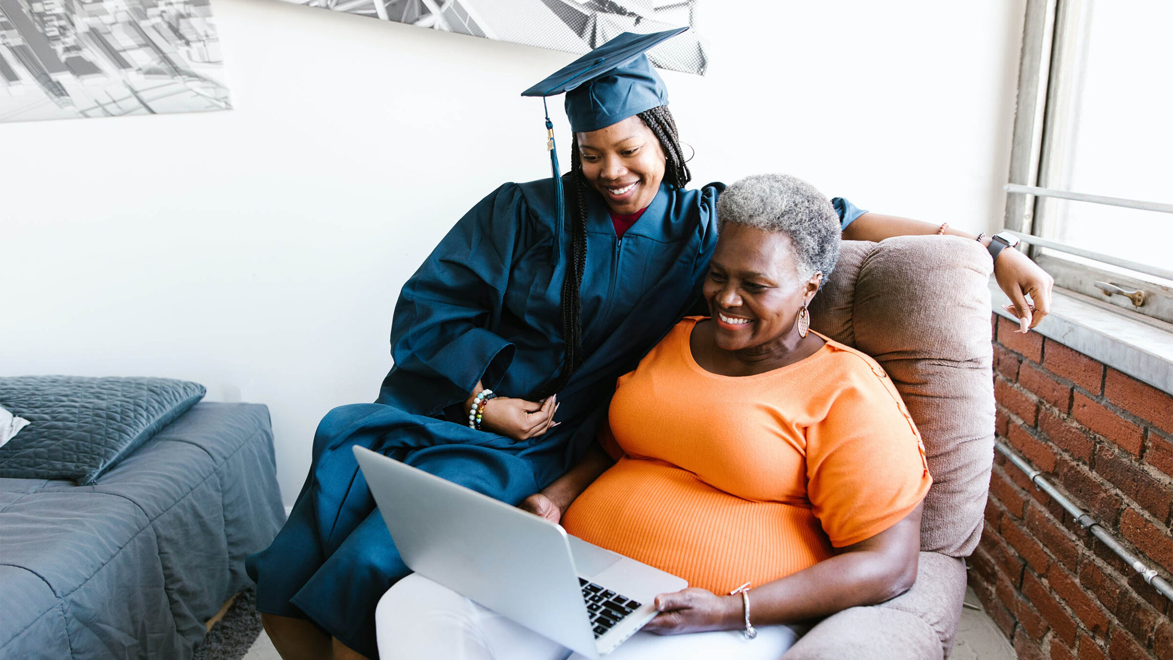 a family looking at a laptop while sitting in a chair. one person wears a bright orange shirt while the other wears a blue cap and gown