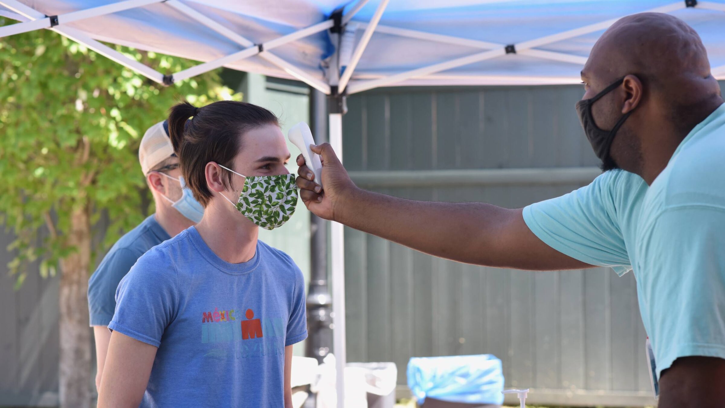a student wearing a mask having their temperature taken on campus by a staff member while wearing a mask