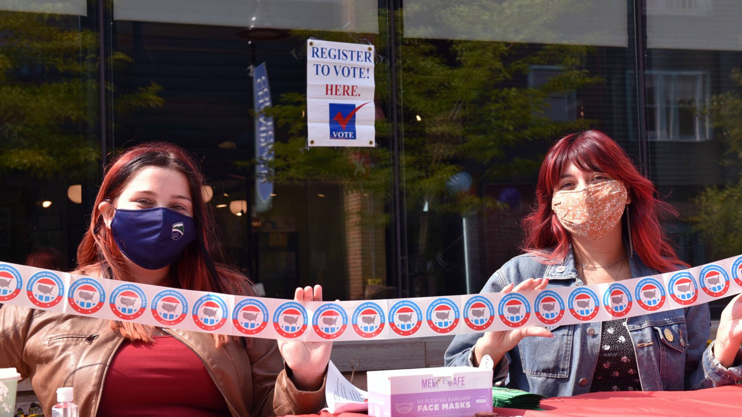 two students wearing masks holding a roll of voting stickers