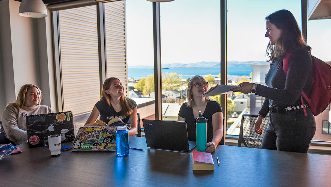 students in a common room in 194 working on laptops together while one holds out papers
