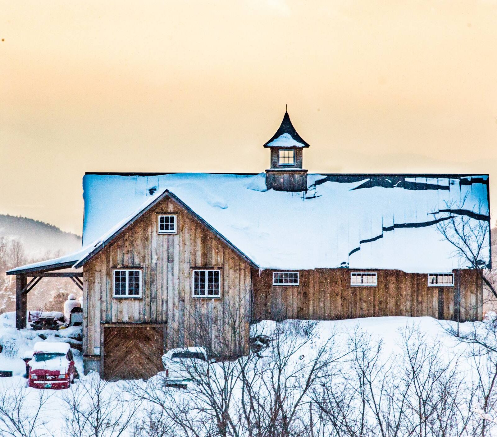 a photo of a snow covered barn in a snowy landscape with a yellow and orange sky