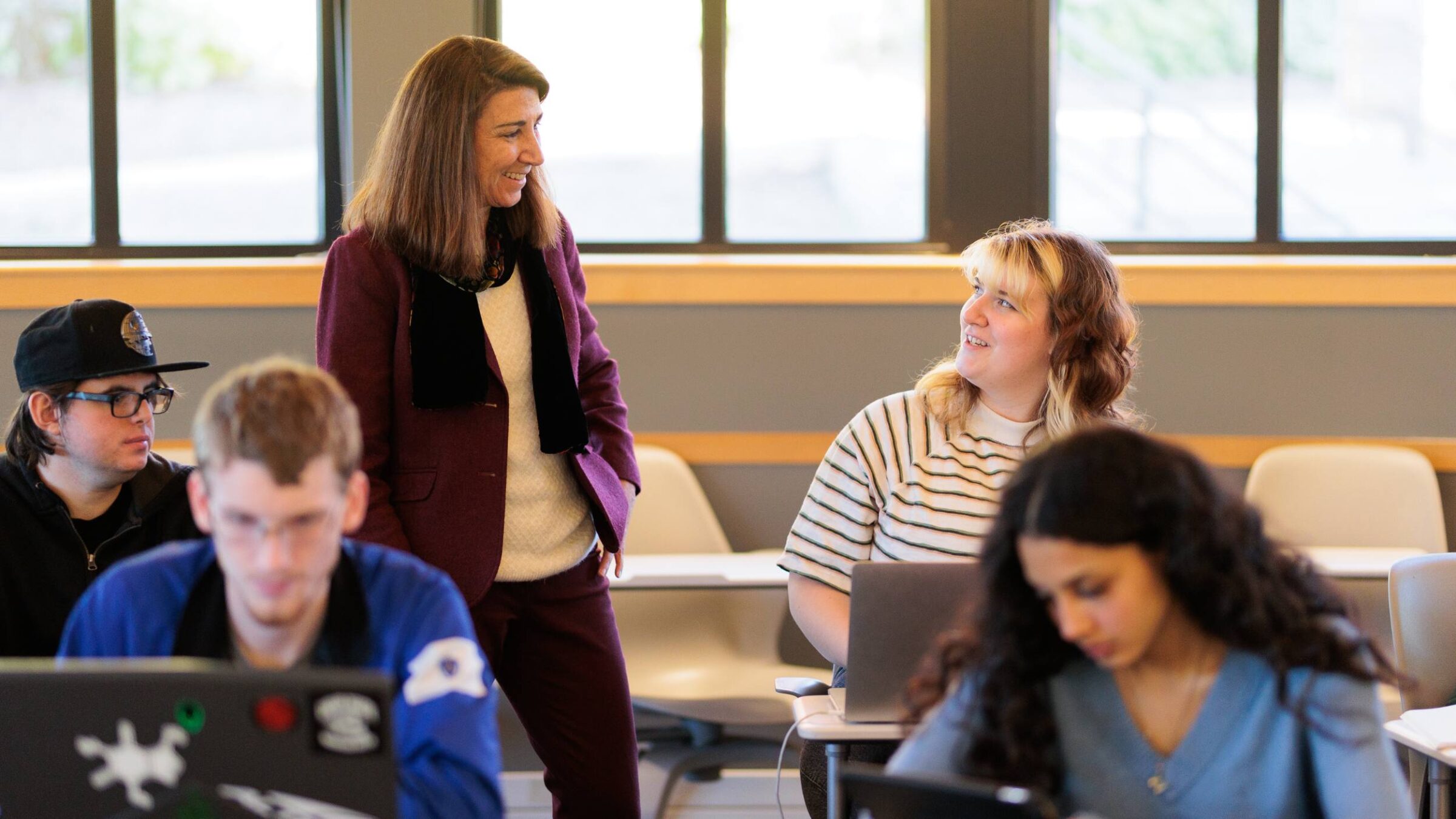 student talking to a professor in a classroom with other students working on laptops and in notebooks