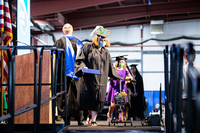 Student walking across the graduation stage with their daughter wearing a cap and gown and holding a diploma