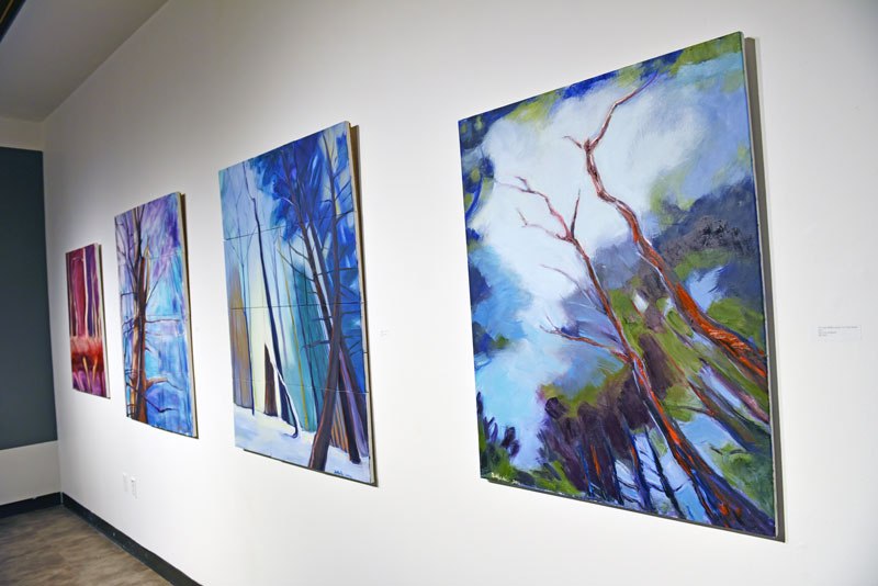 A collection of blue and red paintings on a white wall in Champlain's Art Gallery painted by Ann DeMarle