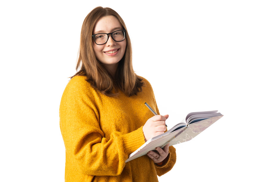 a student wearing a yellow sweater standing in front of a white background writing in a notebook