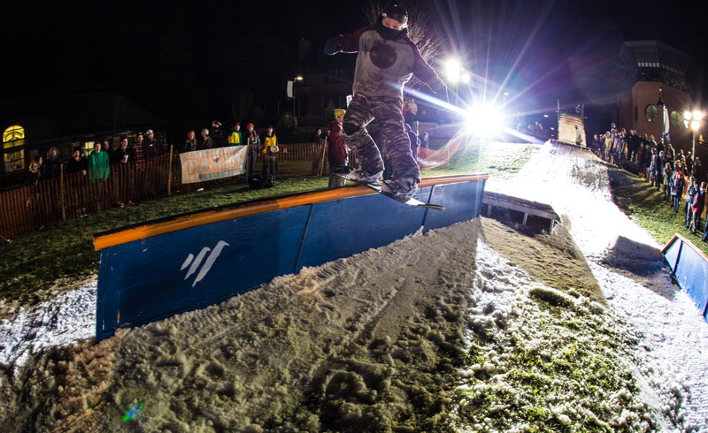 a student riding a snowboard across a rail during the campus rail jam in the quad