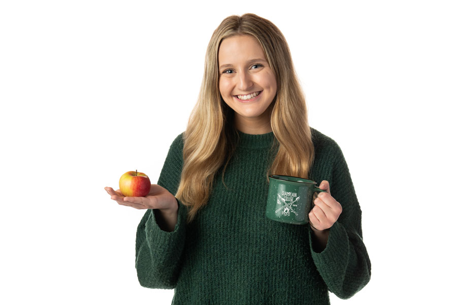 a student wearing a green sweater holding an apple and a green Champlain mug against a white background