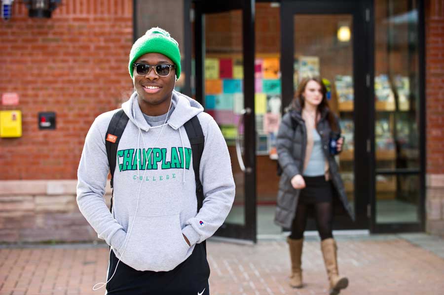 a student wearing a champlain college sweatshirt and a green beanie standing and smiling at the camera while another student walks by