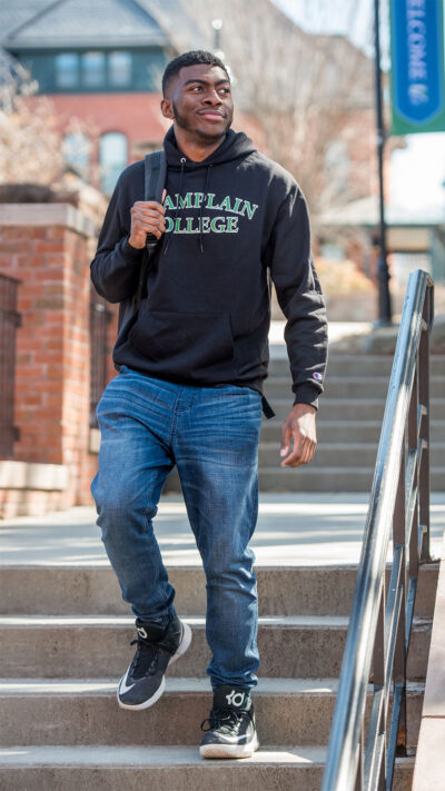 Male student wearing a Champlain College logo sweatshirt walks down a set of steps on campus