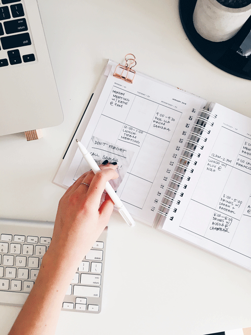 a hand sticking a sticky note into a planner on a white desk with a laptop, keyboard, and pen
