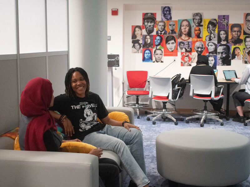 two students sit on a couch in the student resource center space while two people work at a desk in the background