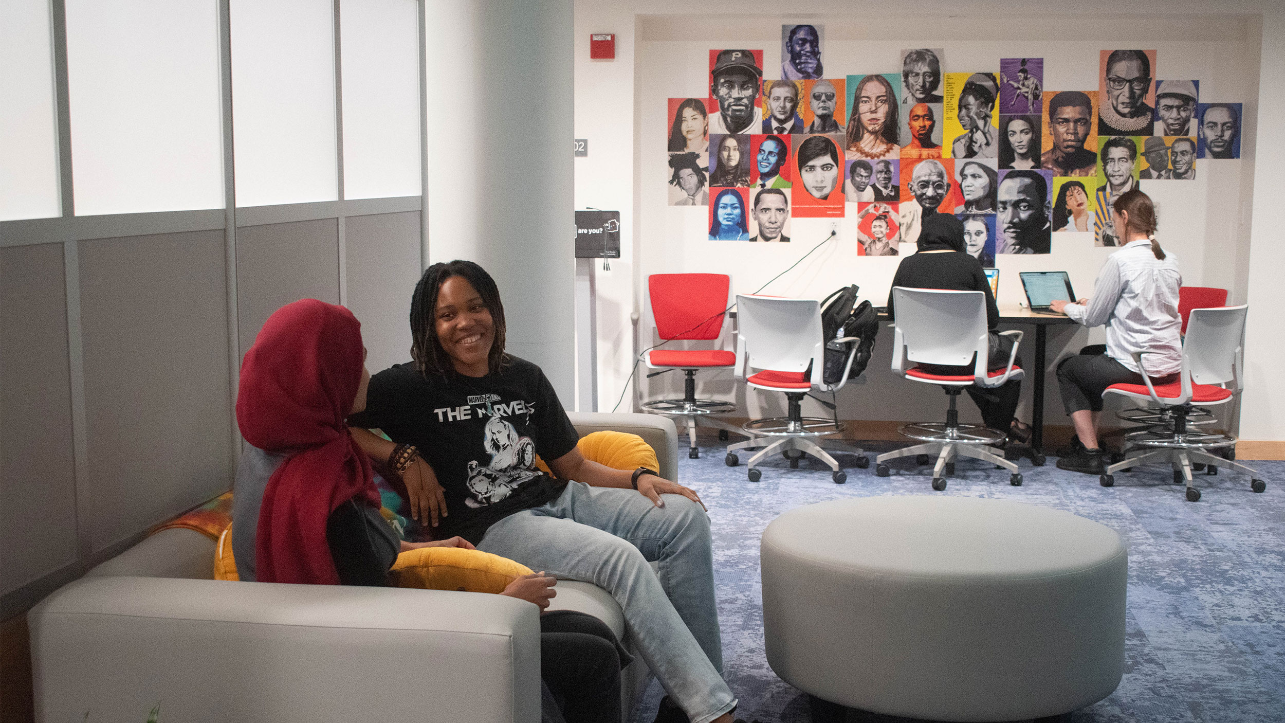 two students sit on a couch in the student resource center space while two people work at a desk in the background