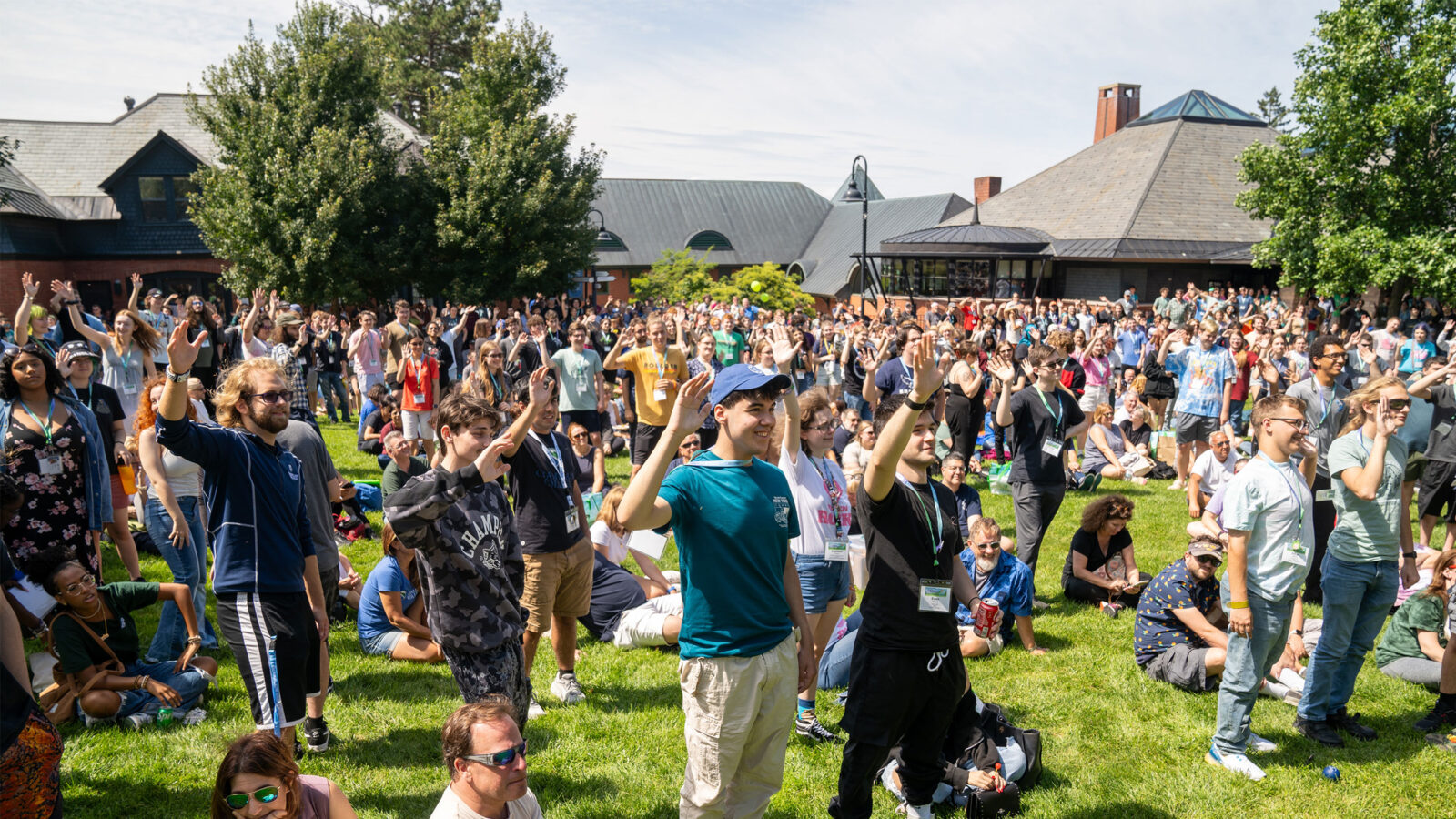 a crowds of hundreds of students and families sit and stand on the lawn, raising their hands