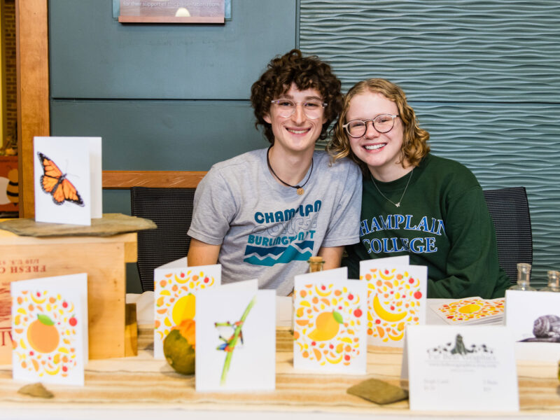 two students smile big sitting at a table covered in homemade cards with yellow fruit drawn on them