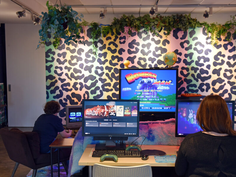 two people play games on a PC and game console in front of a big colorful wall