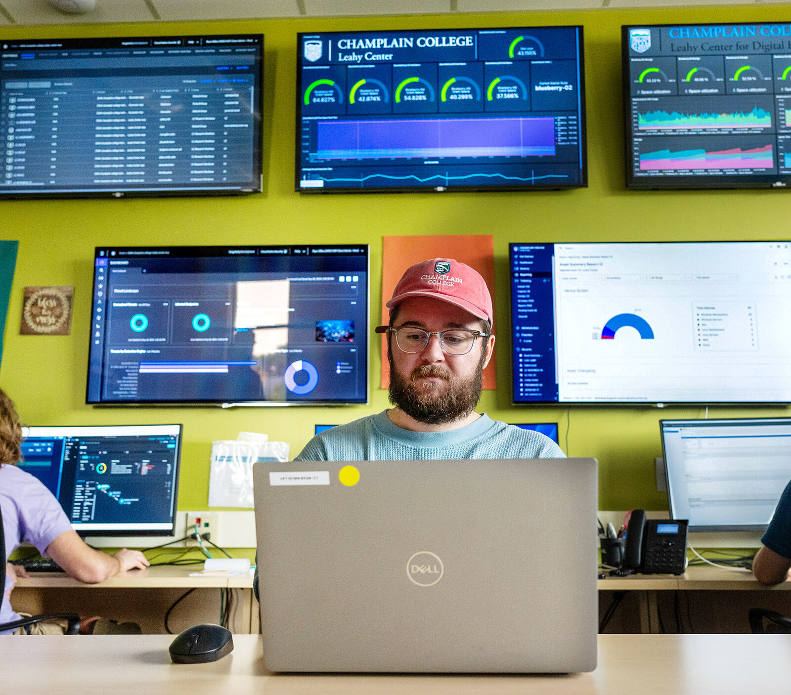 people at computers, large screens showing various data hang on the wall