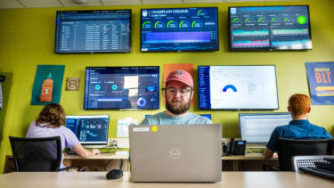 people at computers, large screens showing various data hang on the wall