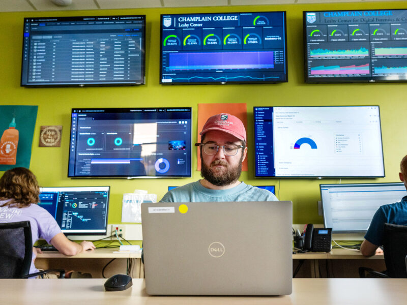 people at computers, large screens showing various data hang on the wall