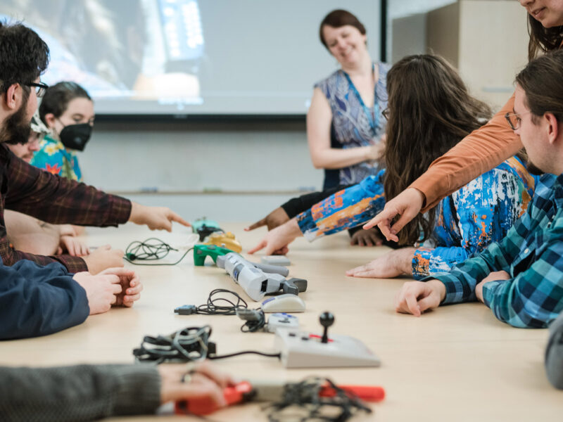 students gathered around a large table where game controllers make a line down the center of the table