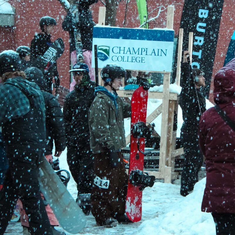 Students standing in the snow near a Champlain College sign holding snowboards