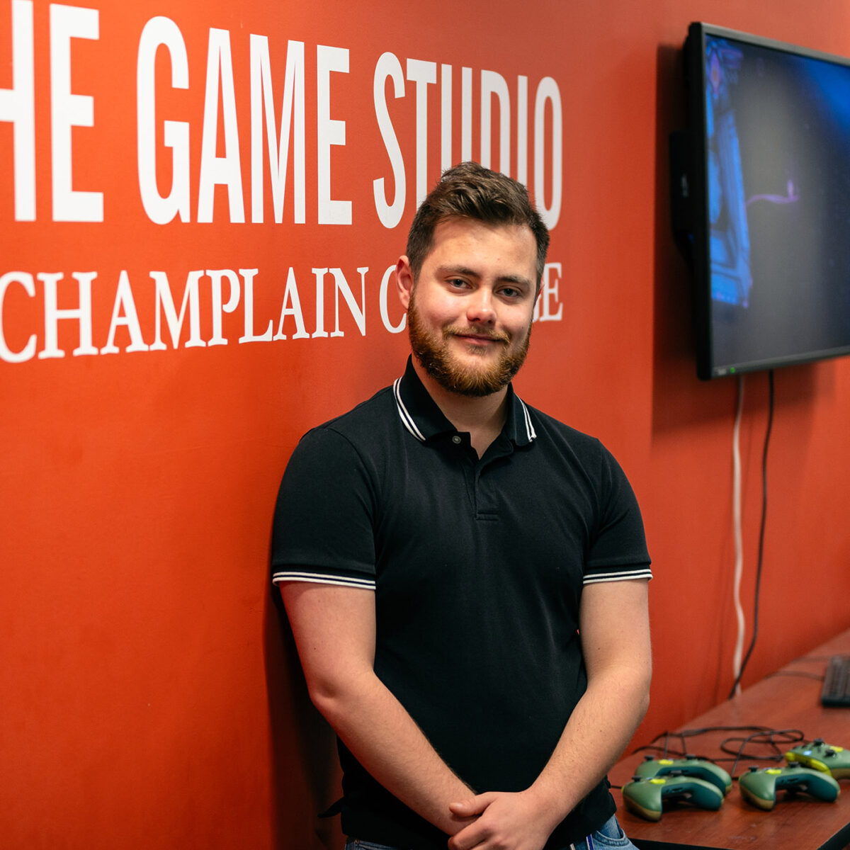 student poses near a sign on the wall that says "The Game Studio at Champlain College"; digital screens and game controllers in background