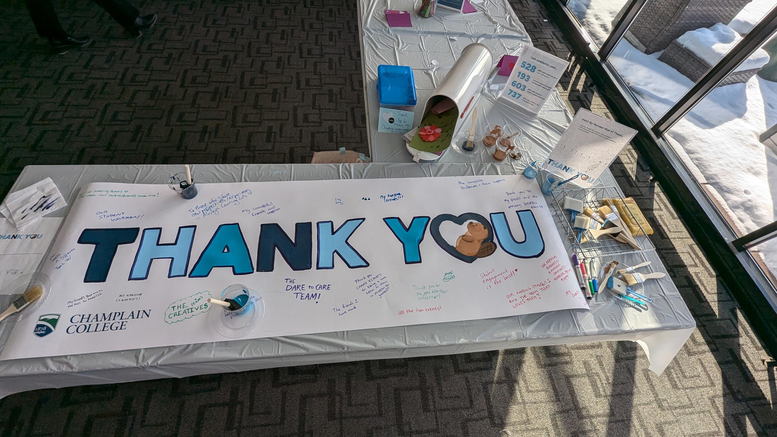 A top-down view of a table with various paints, markers, and signs reading THANK YOU CHAMPLAIN. Various handwritten notes surround a poster on the table.