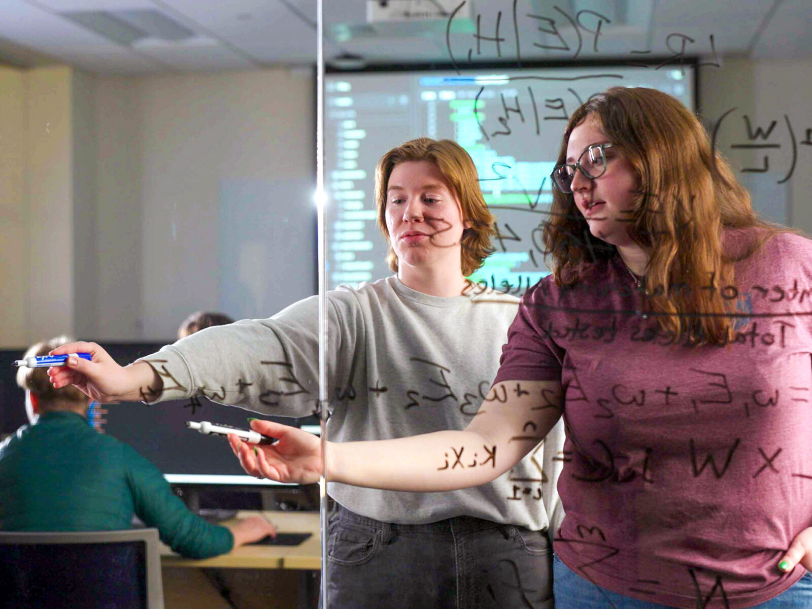 two students write out equations on a glass wall; other students work in background
