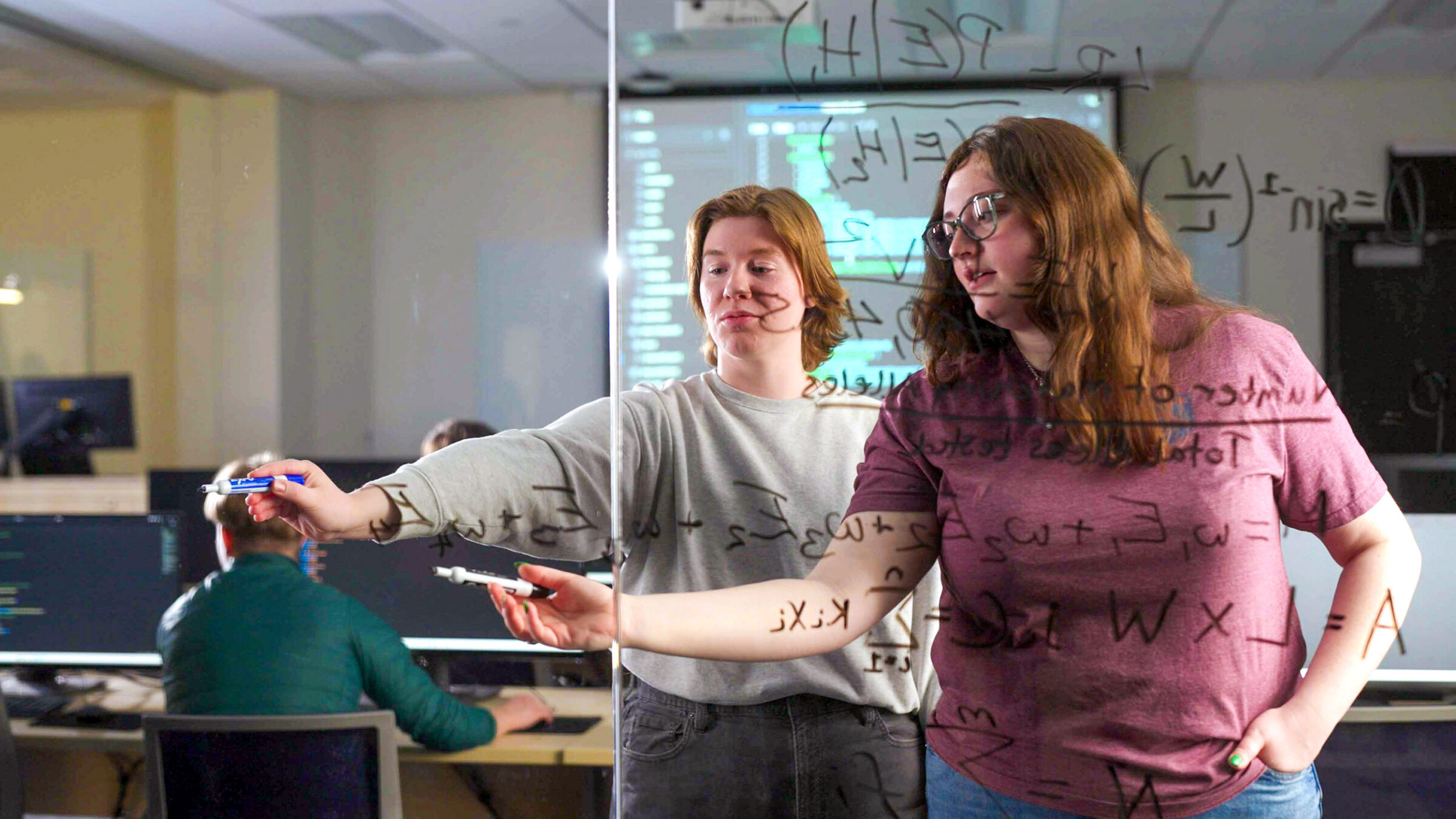 two students write out equations on a glass wall; other students work in background