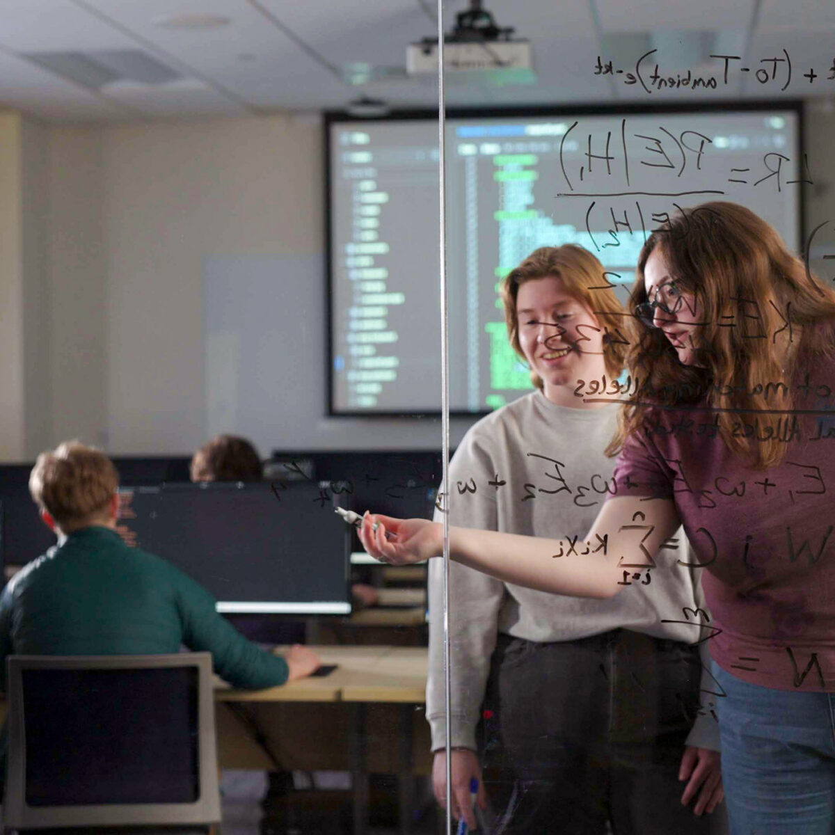 two students write out equations on a glass wall; other students work in background