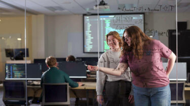 two students write out equations on a glass wall; other students work in background