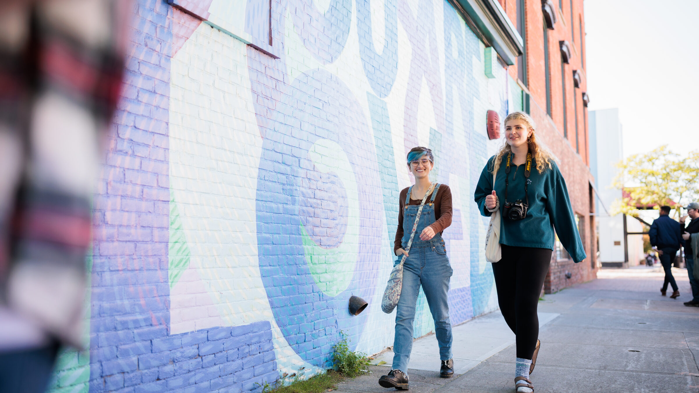 two students talk by an outdoor wall that says "you are loved"