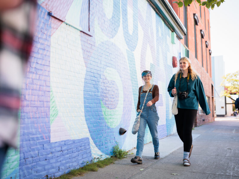 two students talk by an outdoor wall that says "you are loved"