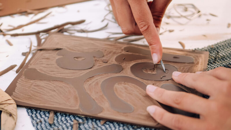 A hand uses a tool to carve into a piece of rubber and create a stamp design.