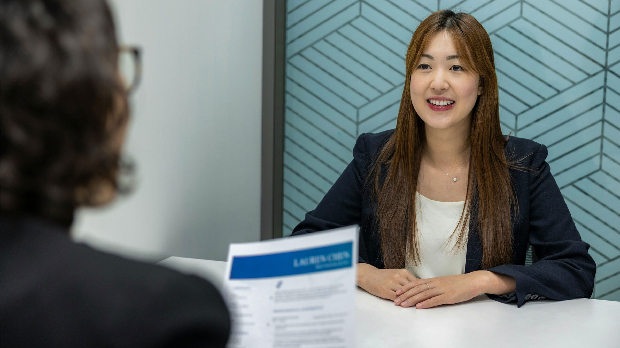 person smiles sitting at a table, sitting across from a person who is reading their resume