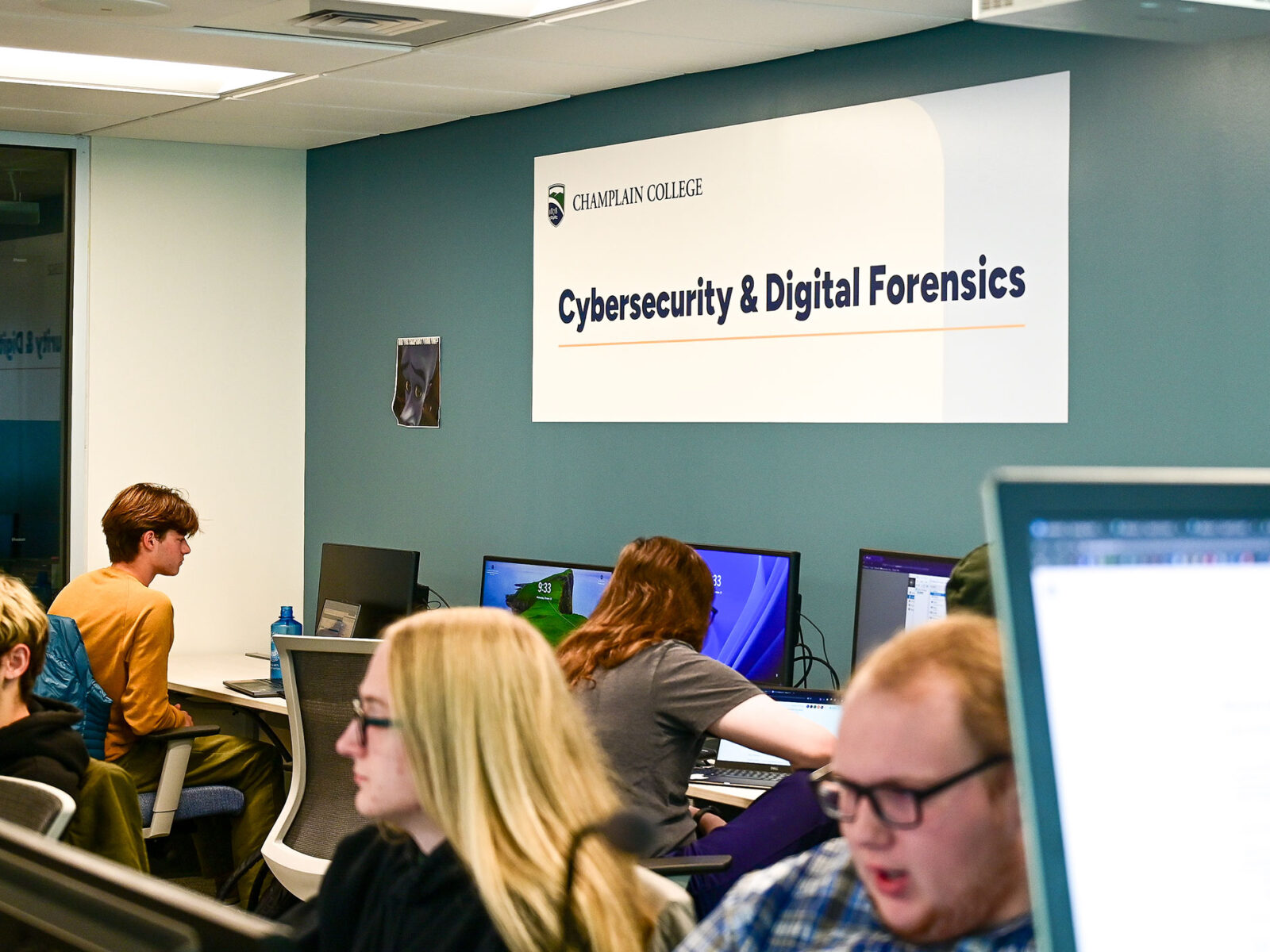 A Champlain College Cybersecurity & Digital Forensics sign hangs in a computer lab.