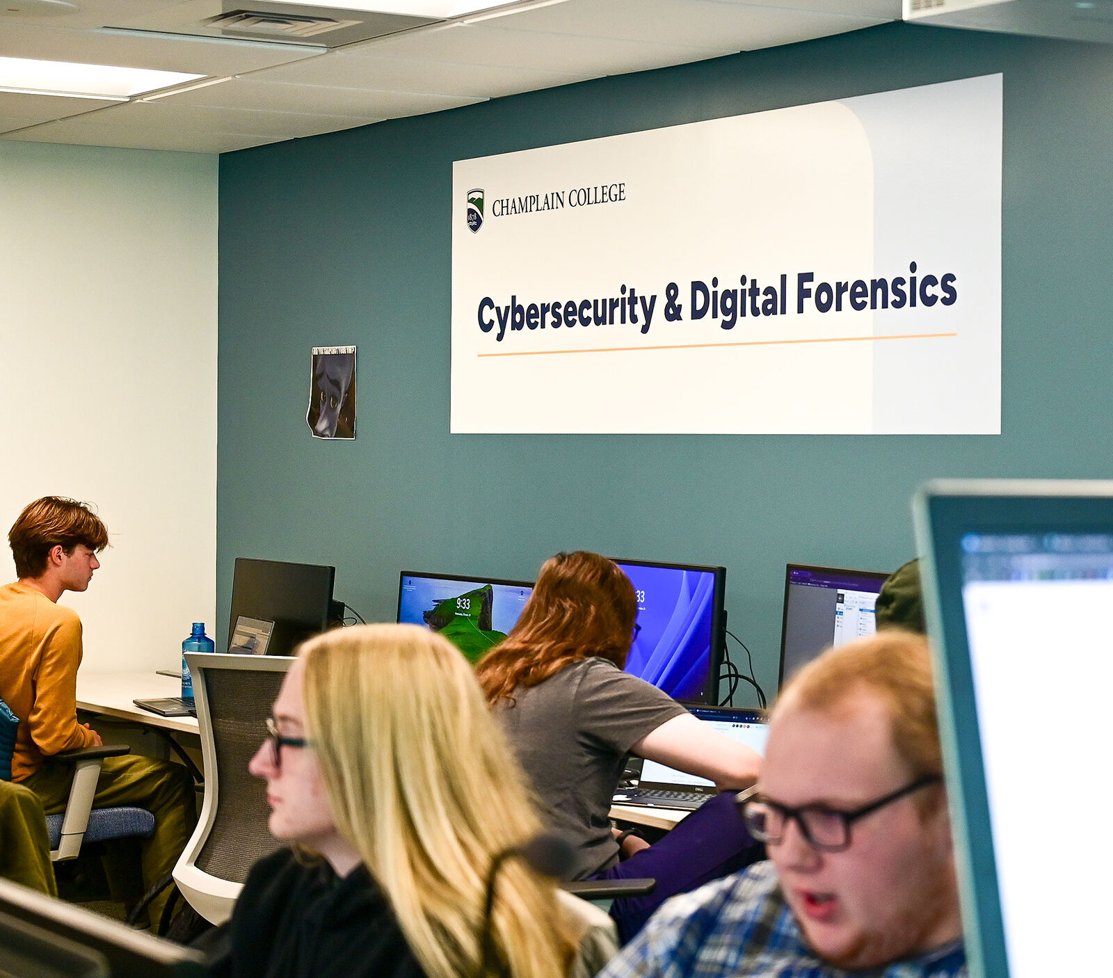 A Champlain College Cybersecurity & Digital Forensics sign hangs in a computer lab.