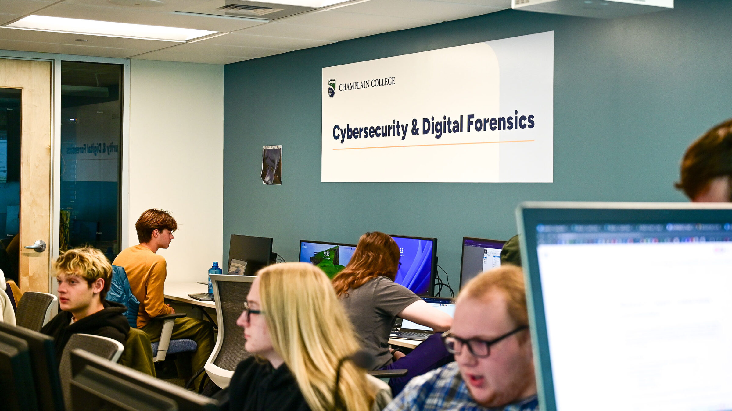 A Champlain College Cybersecurity & Digital Forensics sign hangs in a computer lab.