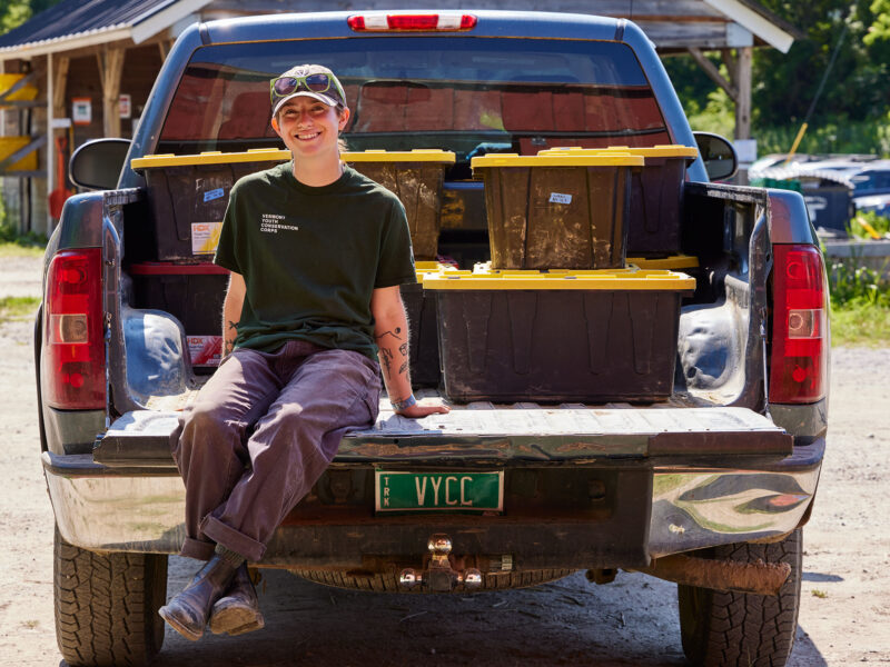 Morgan Chesnais sits on the bed of a pickup truck at their internship at an 11-acre farm owned by VYCC.