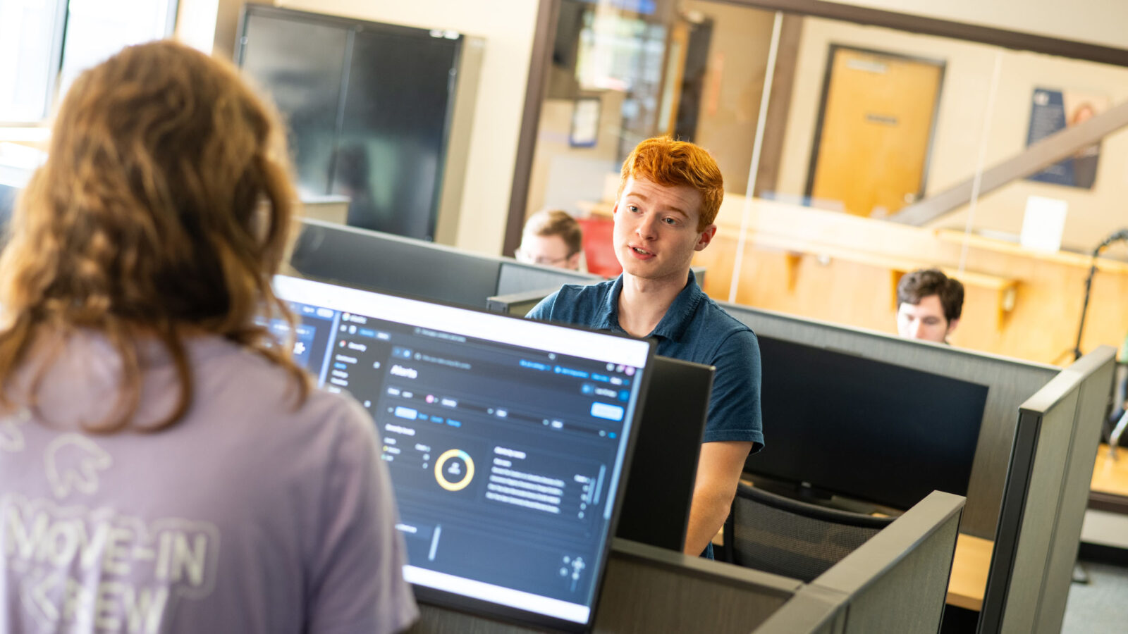 Two students working in the Leahy Center in front of computer screens, having a conversation
