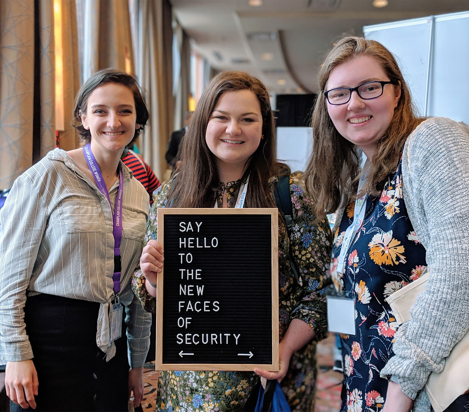 Students at a conference hold a sign that says "Say hello to the new faces of cybersecurity."