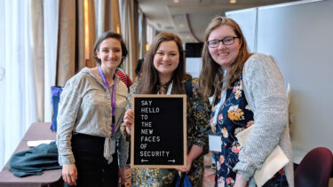 Students at a conference hold a sign that says "Say hello to the new faces of cybersecurity."